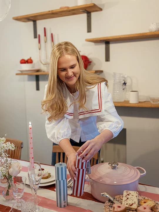 Matilda Bea leaning over a table repositioning a salt cellar. Around her is a pink Le Creuset crock pot, wine glasses and flowers in vases.