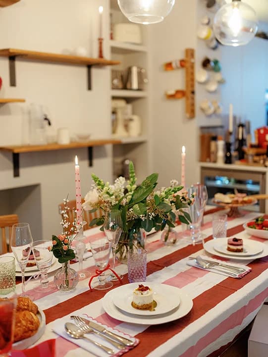 Matilda Bea's tablescape. Stripe tablecloth on a table with glasses, flowers and candles on top. Behind there is a wall with shelves that have pots and candles on them