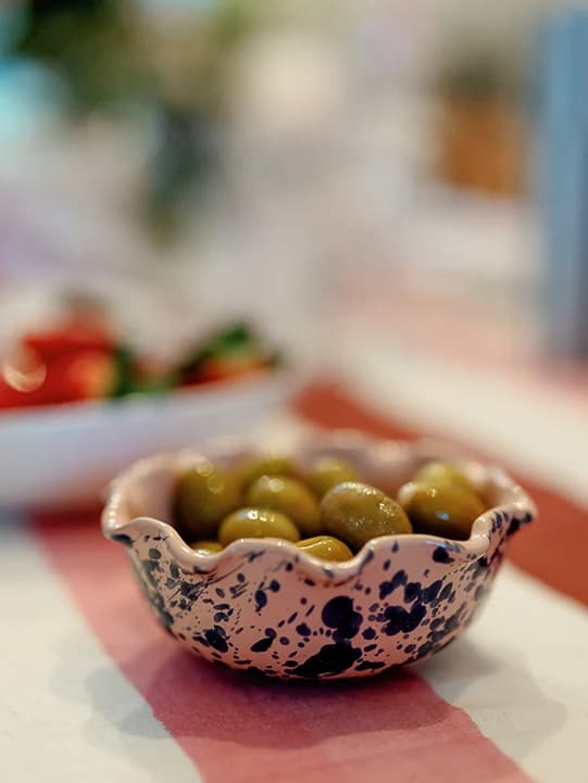 Green olives in a small, splatter bowl on a pink, white and burgundy stripe tablecloth