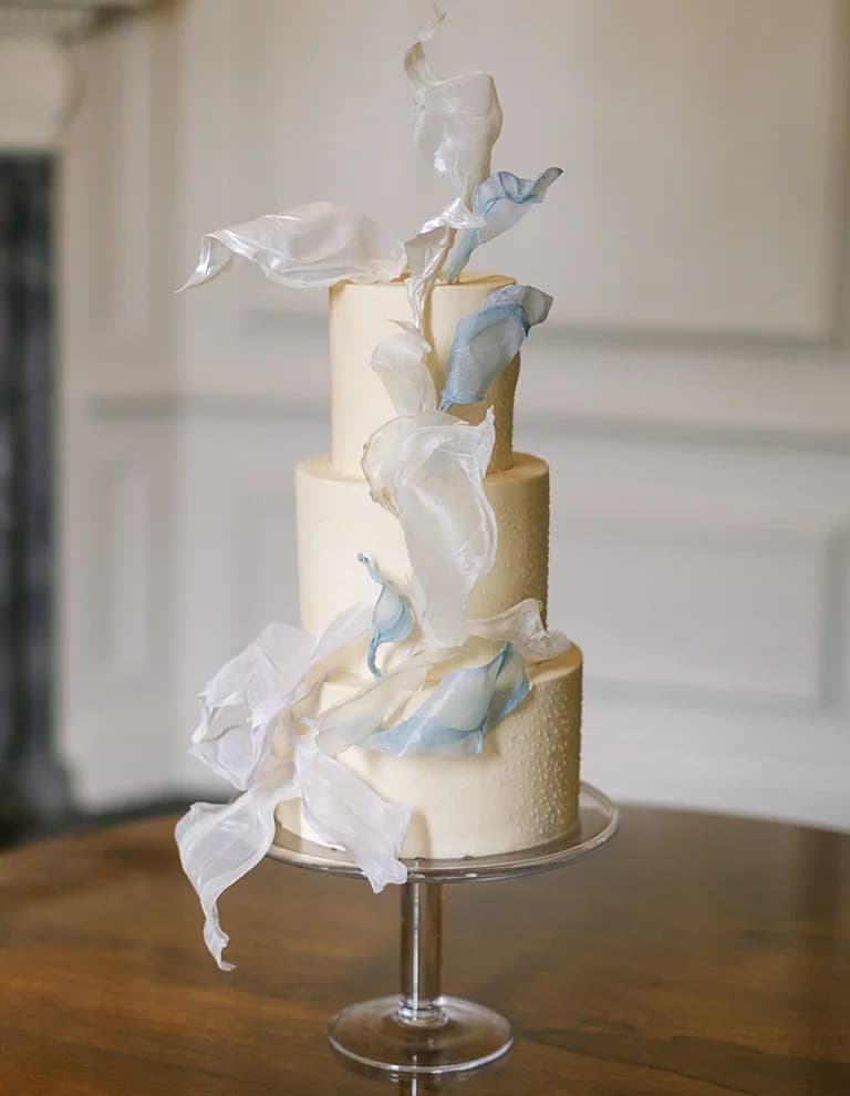 A three tiered wedding cake adorned with pearls and delicate draped fabric in white and pastel blue colour