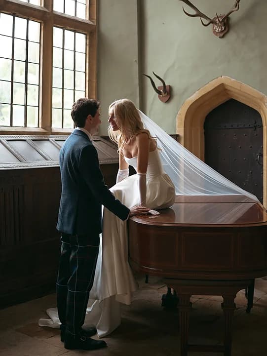 Bride with veil sits on a piano in the Brympton House ballroom, with the groom standing next to her