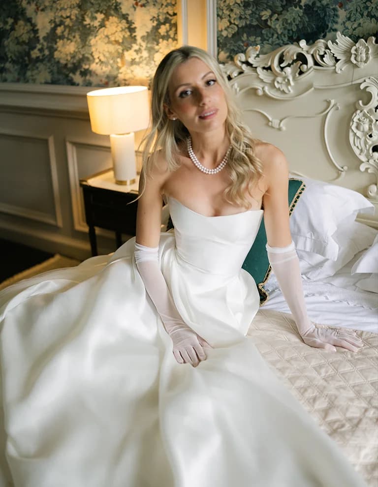 Bride wearing strapless dress and pear necklace sits on a bed in her bridal suite