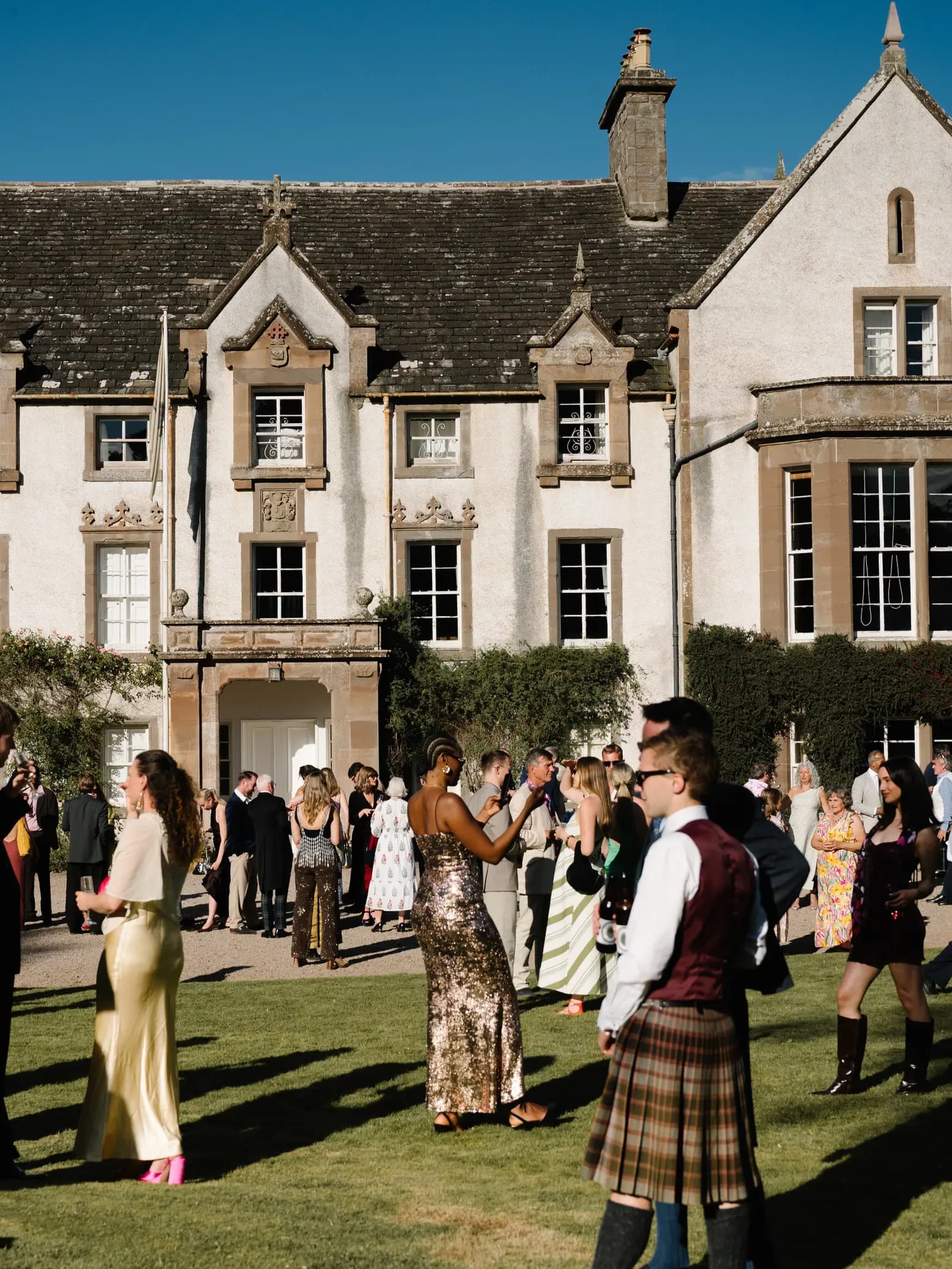 Guests in kilts, sequins and silk dresses mingling on the lawn at Middleton House during a festival wedding in Scotland