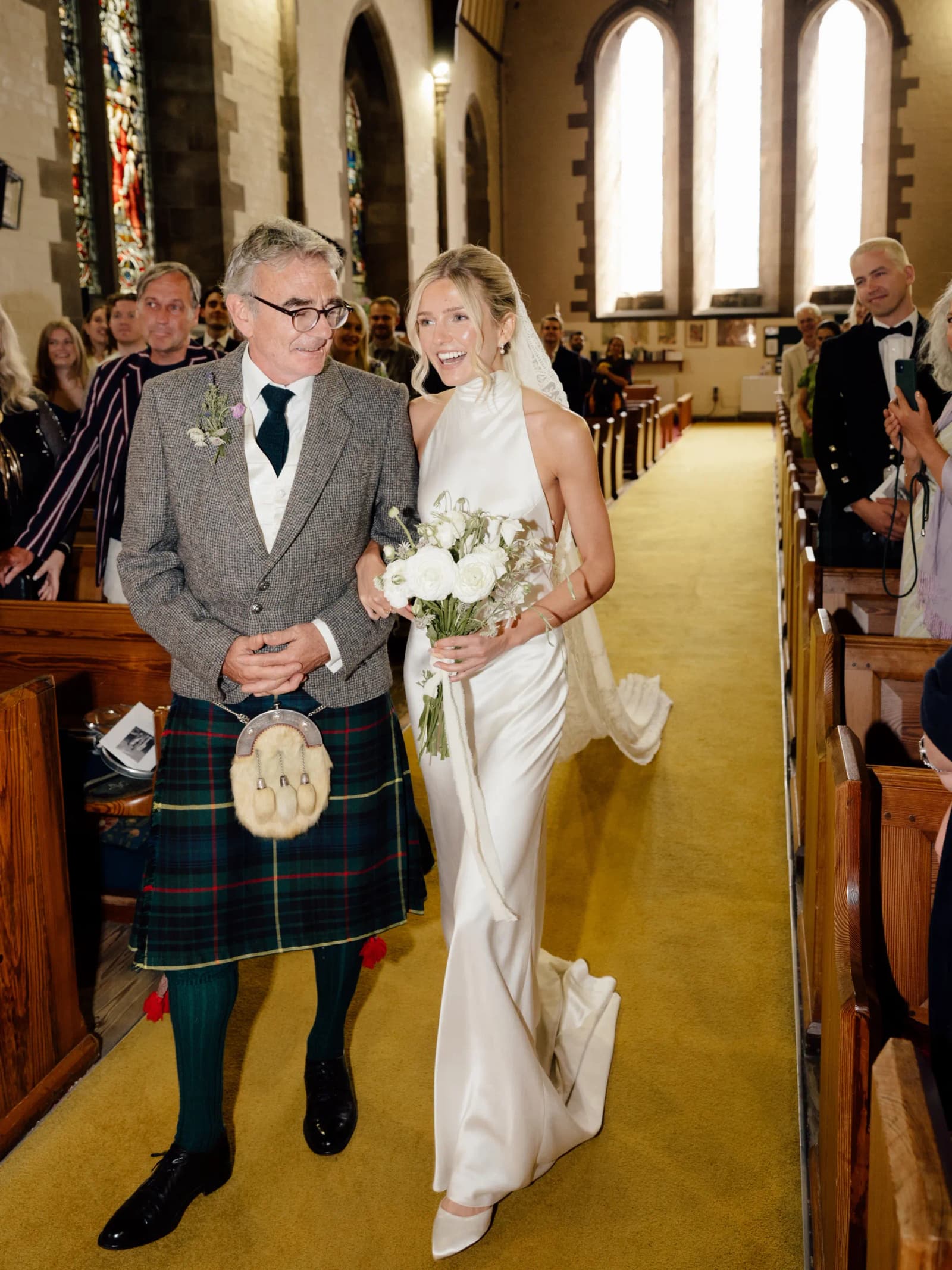 Father of the bride in a tartan kilt walking his smiling daughter down the church aisle at a festival wedding in Scotland