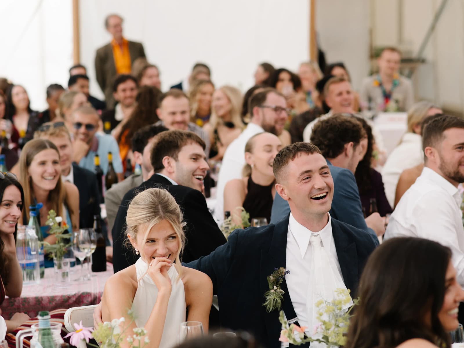 Bride and groom laughing with guests during the wedding speeches in a packed marquee at a festival wedding in Scotland