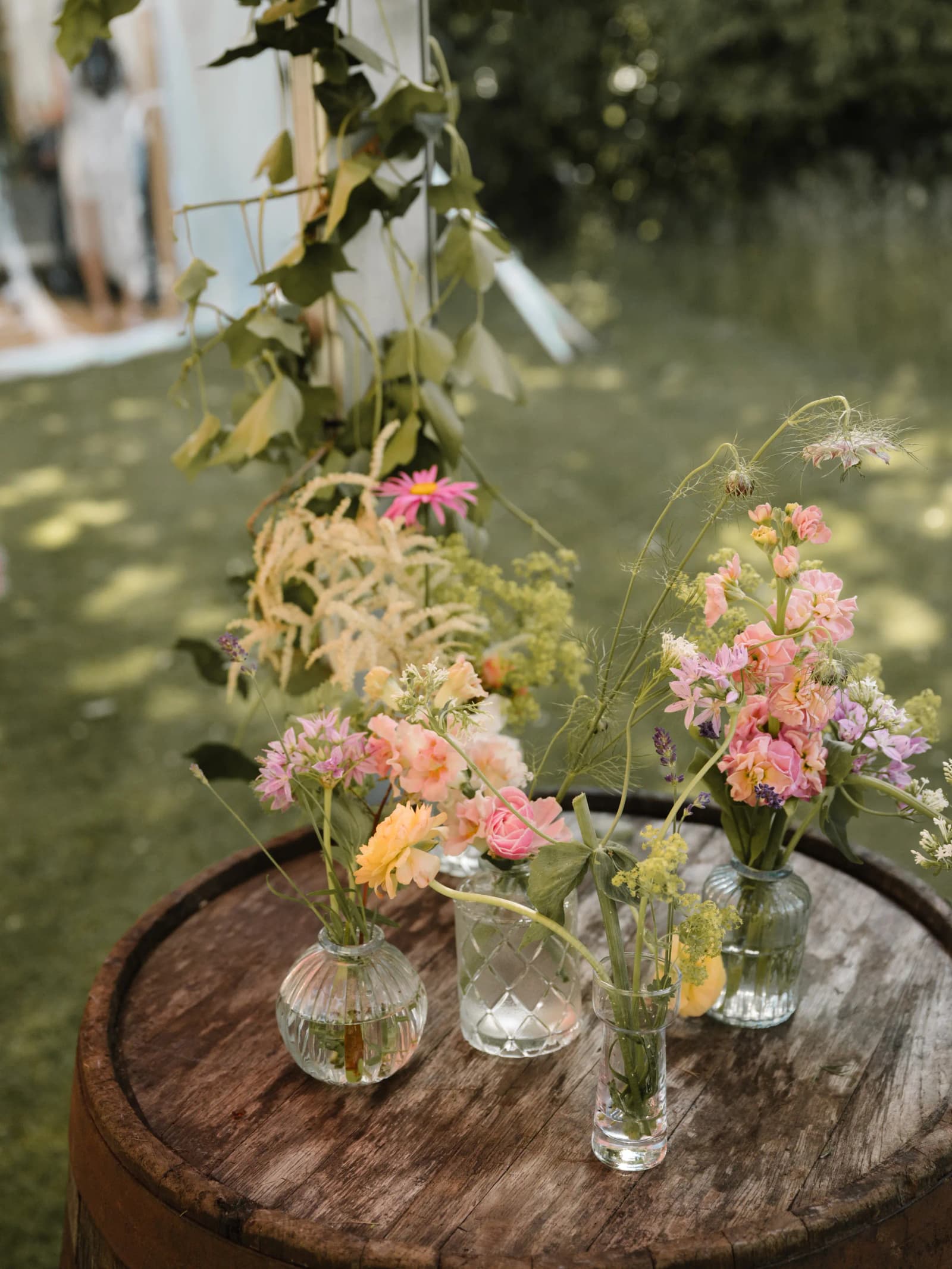 Pink stocks, ranunculus and lady's mantle in vintage glass bud vases on a whisky barrel outside a festival wedding marquee