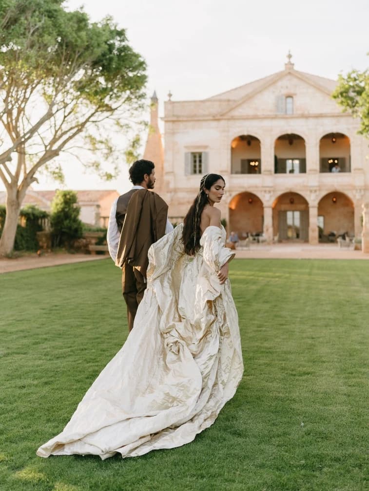 A bride in a flowing ivory gown crosses the lawn with her groom towards a sandstone villa, a romantic backdrop for a Balearic destination wedding.