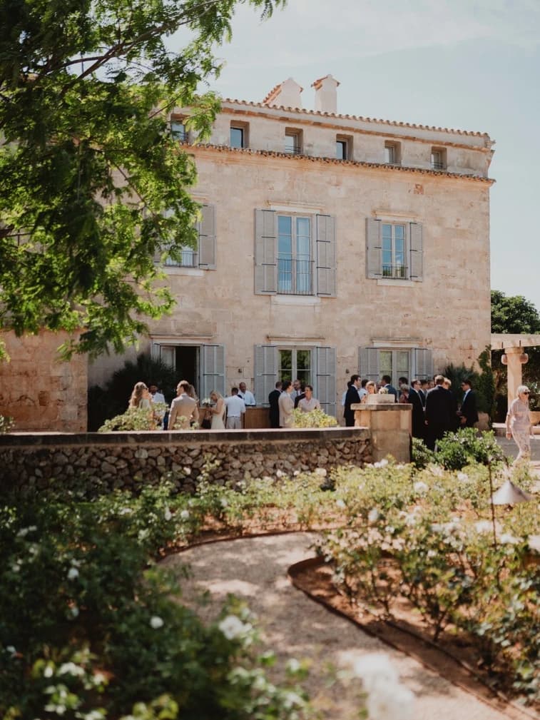 Guests gather for drinks in a rose garden in front of a honey-stone villa, setting a relaxed tone for a destination wedding in the Balearic Islands.